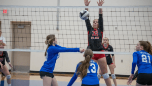 Volleyball match showing a player hitting the ball over the net with a middle blocker attempting to block the hit.