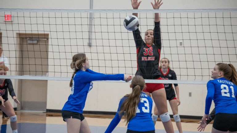 Volleyball match showing a player hitting the ball over the net with a middle blocker attempting to block the hit.