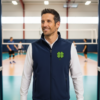 Man wearing navy soft shell vest with clover embroidery inside a gym with volleyball court background
